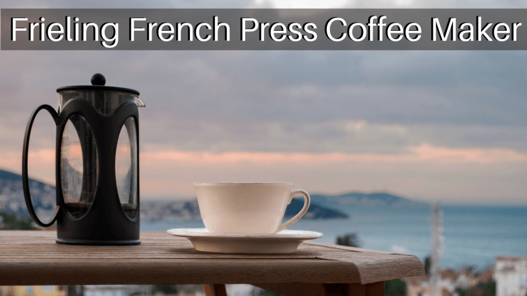 A French press and a white coffee cup and saucer sitting on a wooden table with mountains and the ocean in the background