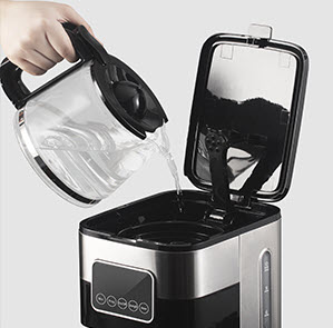 A black handled glass water jug pouring water into a silver and black coffee machine.