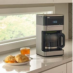 Braun's Brew Sense Drip Coffee Maker 12 Cup Black sitting on a counter top in front of a small glass window with dessert on a plate next to it.