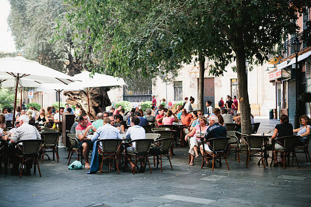 A large crowd of people drinking coffee at an outdoor coffee shop under trees and sun umbrellas showing why is coffee so popular