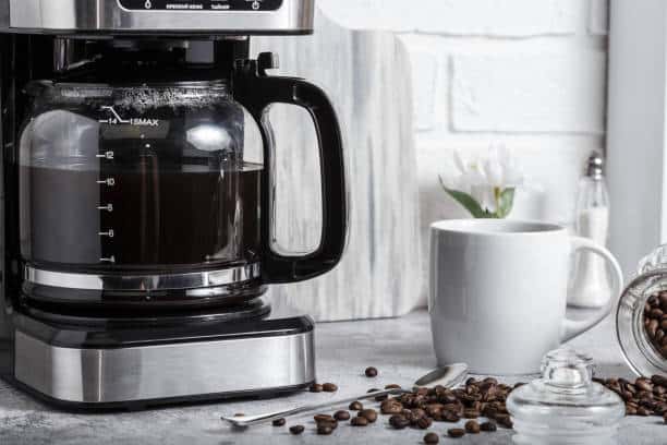 A black electric drip coffee machine with a glass teapot brews a morning drink. Household appliances, a white cup and a jar of beans on the kitchen table on the countertop at home.