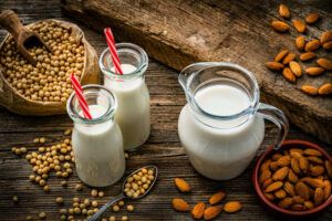 High angle view of a glass bottle and a jar filled with soy milk and almond milk shot on rustic wooden table. Soy beans are around the bottles