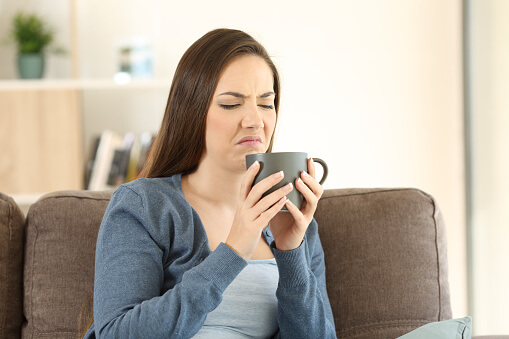 A young woman pulling a face of dislike while drinking her coffee from a mug while sitting on a lounge