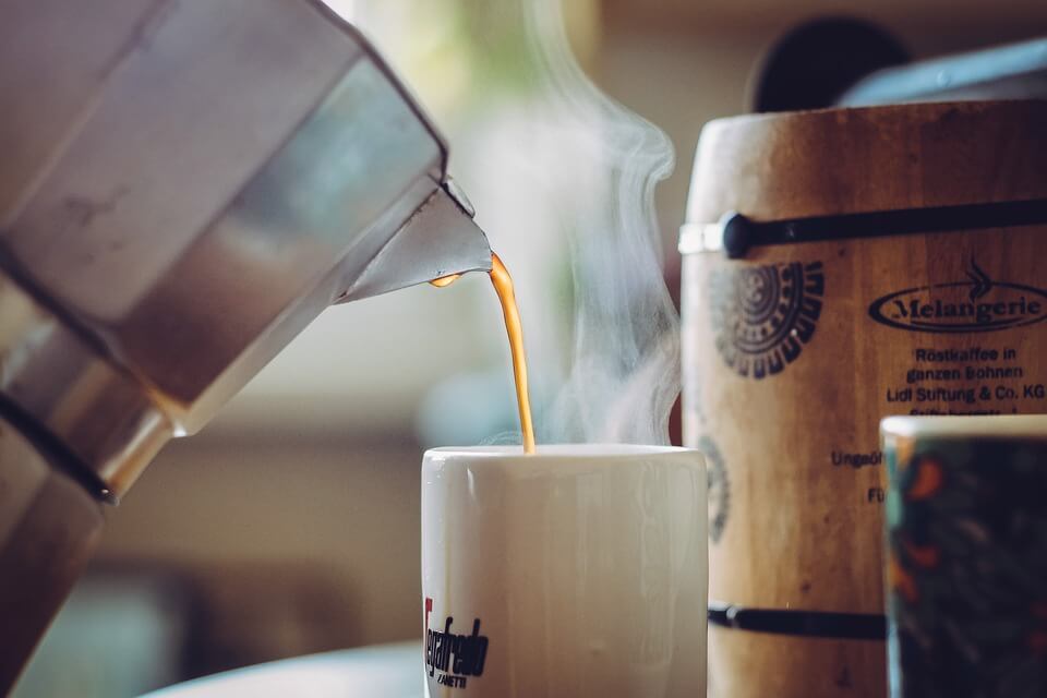 A jug of steaming hot coffee being poured into a white coffee mug with a wooden barrel in the background