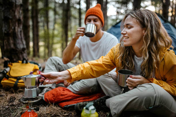 Beautiful and romantic young man and woman preparing hot coffee and tea beverage n portable stove sitting outside tent during hiking adventure, showing how easy instant coffee for campers is to brew.