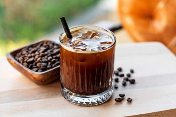 A glass with black coffee, ice cubes and a straw sitting on a table next to a wooden bowl of beans.