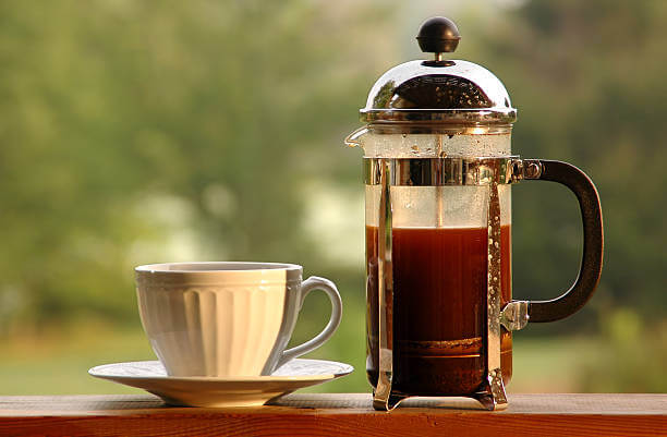 A coffee press and coffee cup shot in the morning light with a lake in the background.