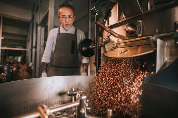 Asian senior man craftsperson observing Freshly roasted coffee beans being removed from the roaster into the cooling cylinder with motion blur on the bean
