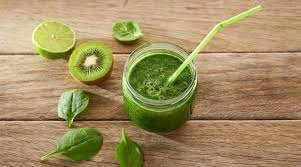 A glass of green vegetable juice surrounded by a kiwi fruit, lime and leaves.