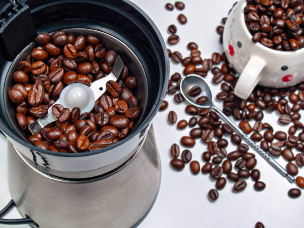 Electric coffee grinder with roasted coffee beans inside and coffee beans scattered on a table with a metal spoon from above and a cup filled with coffee grains shot close-up view from above
