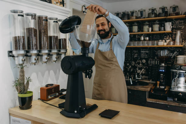 Day in coffee shop. An ethnic man pouring coffee beans into a large coffee grinder