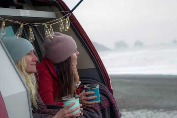 Two young ladies in the back of a car laying down looking out at the ocean with travel coffee mugs in their hands.