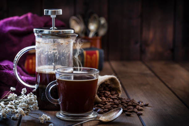 Front view of a transparent glass coffee cup and a coffee french press placed at the left of a rustic wooden table. A burlap sack with roasted coffee beans and old books at background.