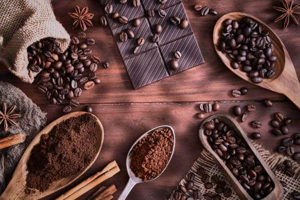 Coffee beans, spices and chocolate sitting in wooden spoons on a wooden table top. Top view.