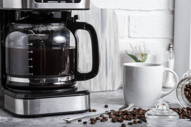 A black drip electric coffee machine with a glass teapot brews a morning drink. Household appliances, a white cup and a jar of beans on the kitchen table on the countertop at home.