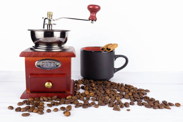 Wooden coffee grinder and black mug with cinnamon sticks on scattered coffee beans on white background