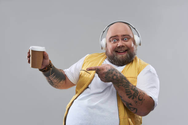 Excited overweight bearded man wearing big headphones while standing isolated on the grey background and pointing to the cup of coffee in his hands