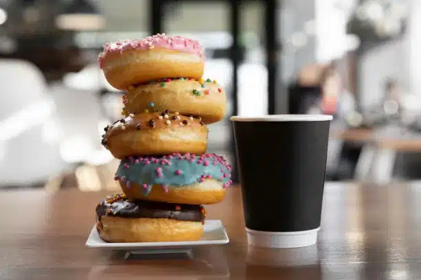 Colorful glazed coffee and donuts pyramid on a white plate standing on a wooden table close to black desposable paper coffee cup