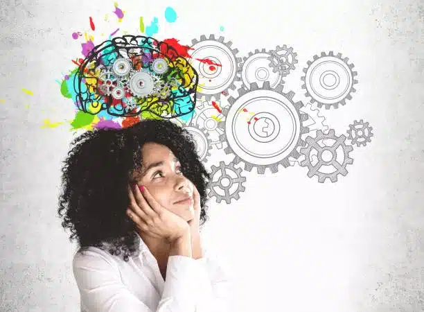 Smiling young African American woman in white shirt looking at colorful brain sketch with gears drawn on concrete wall. Concept of brainstorming